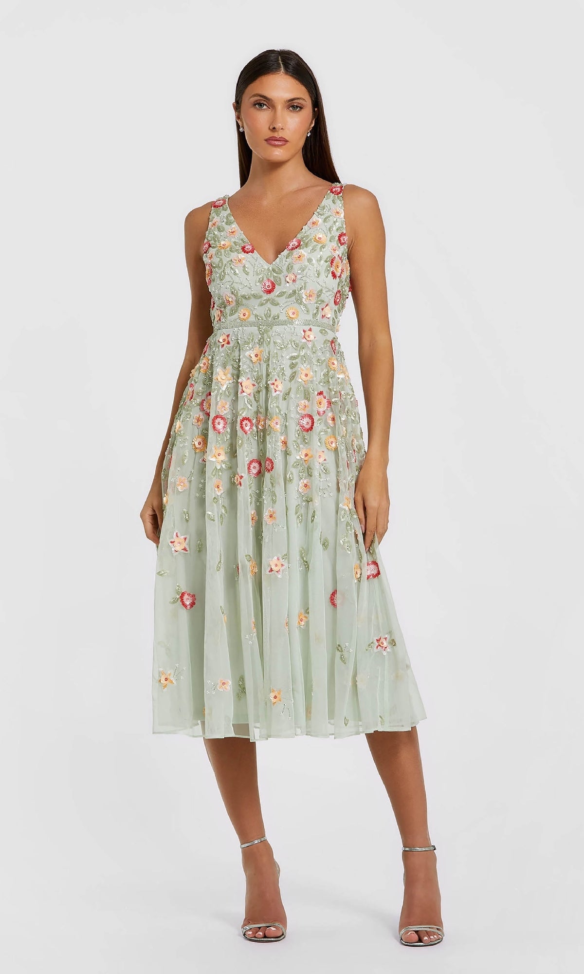 A woman in the Mac Duggal 5527 Floral-Embroidered Party Dress—sleeveless, light green, tea-length with multicolored floral embroidery—and silver heels stands against a plain background.
